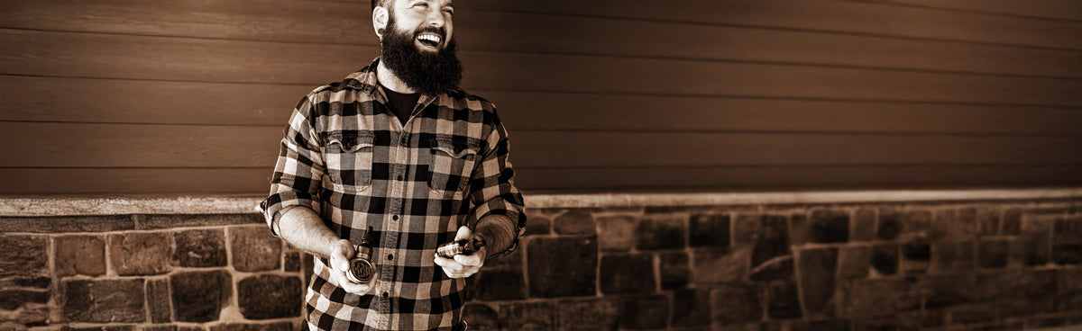 Man with a beard wearing a plaid shirt standing against a brick wall.