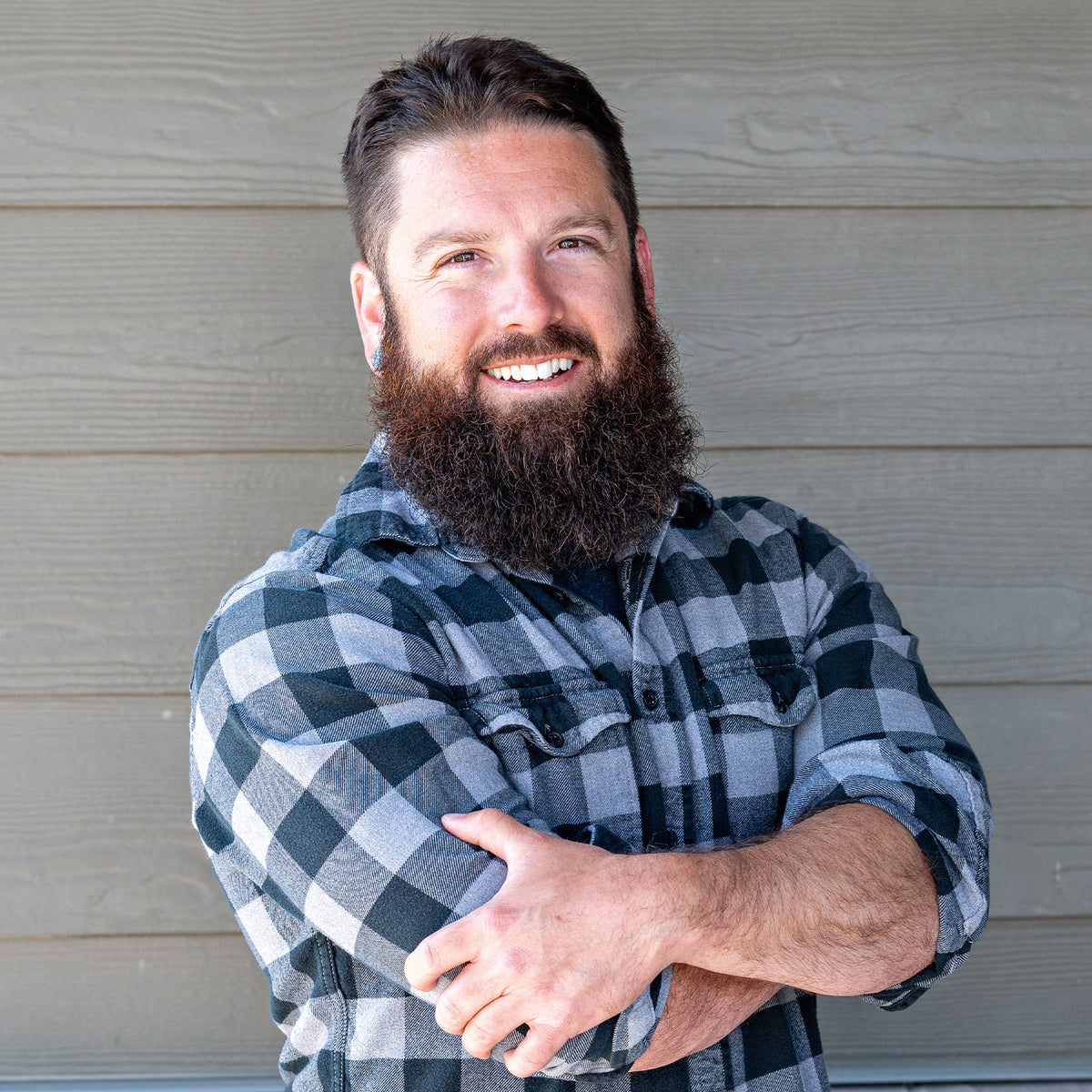 Man with a beard wearing a plaid shirt standing against a neutral wall.